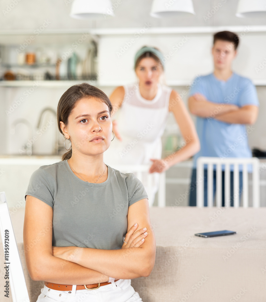 Obraz premium Sad offended young woman sitting at kitchen table during argument with dissatisfied husband and mother-in-law standing behind on blurred background. Concept of family fights