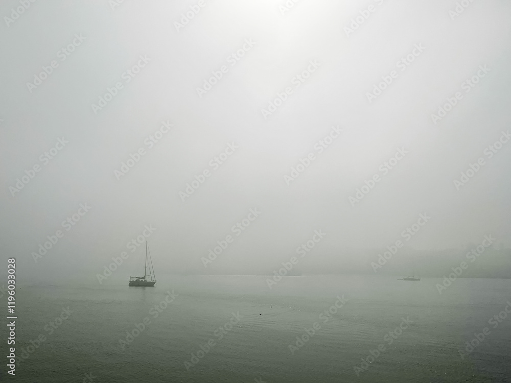 Fototapeta premium A tranquil scene of a sailboat drifting on calm waters, surrounded by dense fog that obscures the horizon. The minimalist composition and soft tones evoke feelings of solitude, serenity, and mystery