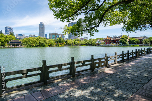 The waterfront of Moon Lake Park in Ningbo, Zhejiang, China. A paved pathway winds along the lakeshore with traditional stone railing and a modern skyline in the distance. A Chinese urban parkland.