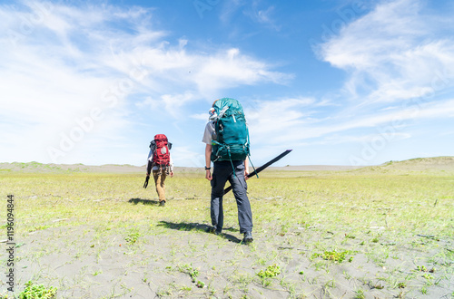 Two people with backpacks trekking across scenic sand dunes, enjoying the adventure and connection with nature under an open sky.