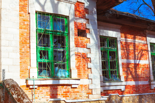 Section of old brick building with green framed windows and broken glass and metal bars. Building is abandoned with shadows of tree branches cast on wall, adding to overall atmosphere of neglect photo