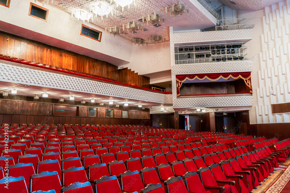 Naklejka premium This image showcases a grand theater interior featuring rows of red velvet seats facing the stage. A blend of modern and classical elements is visible in the rich wood paneling, ornate balconies