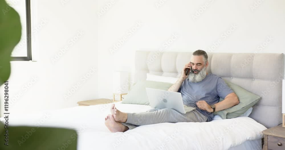 Working from home, man using laptop and making phone call in bedroom, copy space Working from home, man using laptop and making phone call in bedroom, copy space