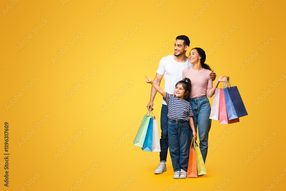 Family Shopping. Cute Little Girl Pointing Finger Aside Showing Something To Her Arab Parents While They Standing Together With Shopper Bags Over Yellow Studio Background, Panorama With Copy Space Family Shopping. Cute Little Girl Pointing Finger Aside Showing Something To Her Arab Parents While They Standing Together With Shopper Bags Over Yellow Studio Background, Panorama With Copy Space
