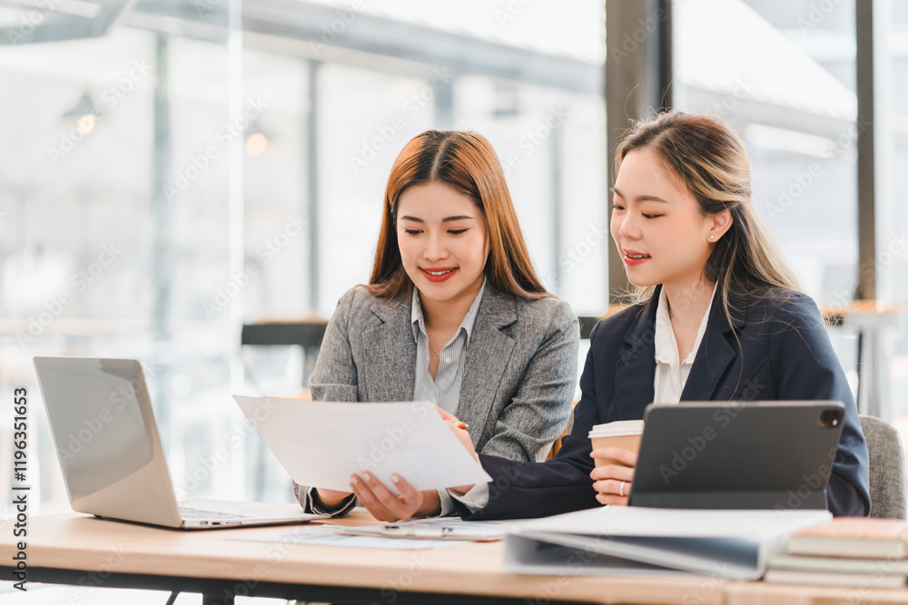 Two businesswomen in formal attire discussing documents at modern office table with laptop, tablet, and coffee cup, showcasing teamwork and collaboration in professional setting Two businesswomen in formal attire discussing documents at modern office table with laptop, tablet, and coffee cup, showcasing teamwork and collaboration in professional setting