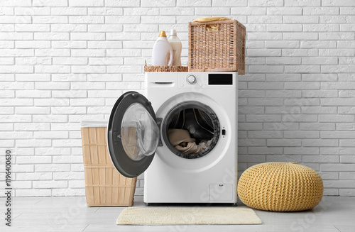 Modern washing machine with laundry, baskets and detergents near white brick wall photo