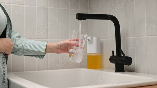 An unrecognizable woman pours tap water into a glass tumbler in the kitchen