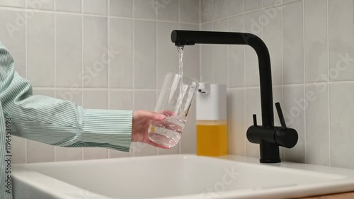 An unrecognizable woman pours tap water into a glass tumbler in the kitchen