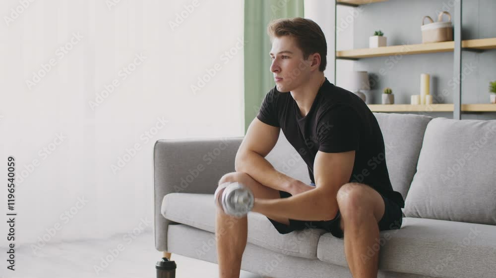 A young and fit man pauses while exercising with a dumbbell at home. He enjoys a healthy lifestyle, emphasizing fitness and well-being in his modern living space. A young and fit man pauses while exercising with a dumbbell at home. He enjoys a healthy lifestyle, emphasizing fitness and well-being in his modern living space.
