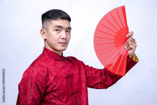 An Asian man in a red traditional Chinese Tang suit holds a red hand fan raised beside his face with a confident expression, cultural heritage, isolated against a plain white background photo