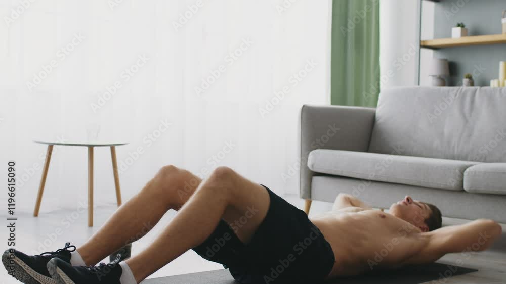 A sporty young man performs exercises on a mat in his living room, focusing on fitness and well-being. His dedication to a healthy lifestyle is evident in his workout routine. A sporty young man performs exercises on a mat in his living room, focusing on fitness and well-being. His dedication to a healthy lifestyle is evident in his workout routine.