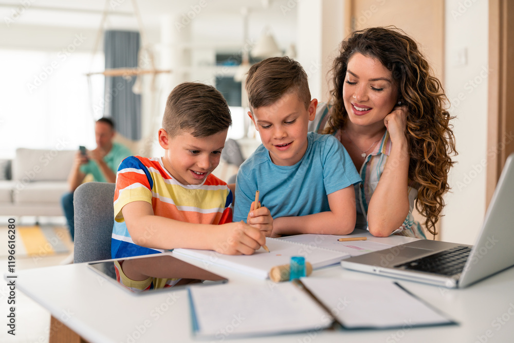 Fototapeta premium Caucasian mother with curly hair helps two elementary-aged sons with homework using laptop and papers at wooden dining table in bright home setting.