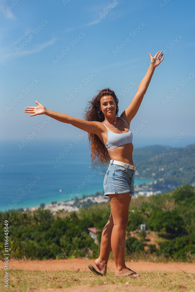A woman stands on a grassy hilltop with arms outstretched, overlooking a vibrant blue ocean, green hills, and a small settlement in tropical Thailand. A woman stands on a grassy hilltop with arms outstretched, overlooking a vibrant blue ocean, green hills, and a small settlement in tropical Thailand.
