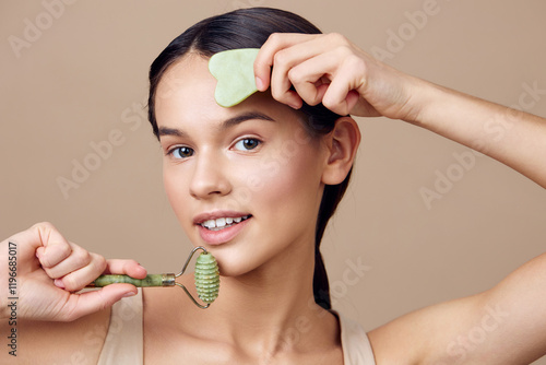 Young woman enjoying a skincare routine with a jade roller and gua sha tool, showcasing healthy skin and relaxation in a serene beige background photo