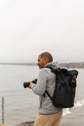 A man with a backpack is standing on a beach, holding a camera photo