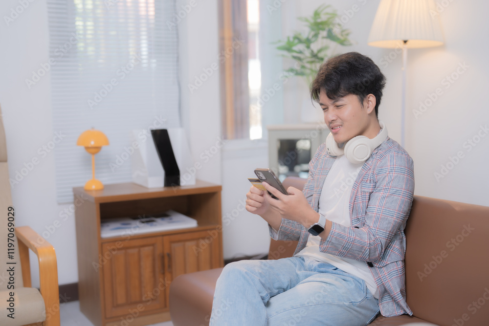 Happy Asian male freelancer sitting on a comfortable sofa at home, holding a credit card and using a smartphone for online shopping, enjoying a seamless e-commerce experience Happy Asian male freelancer sitting on a comfortable sofa at home, holding a credit card and using a smartphone for online shopping, enjoying a seamless e-commerce experience