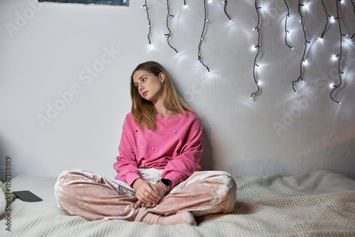 A teenage girl sits pensively on her bed, surrounded by fairy lights, reflecting on her day. photo