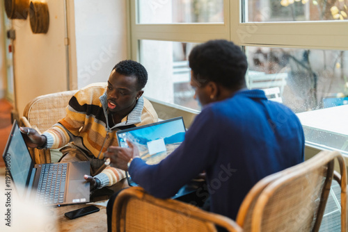 Two young black men actively collaborating on laptops while seated on wicker chairs near large windows in a modern cafe, engaged in an animated exchange of ideas with expressive gestures. photo