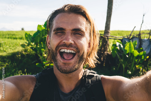 Happy young male tourist with backpack taking selfie against countryside scenery #1197218616