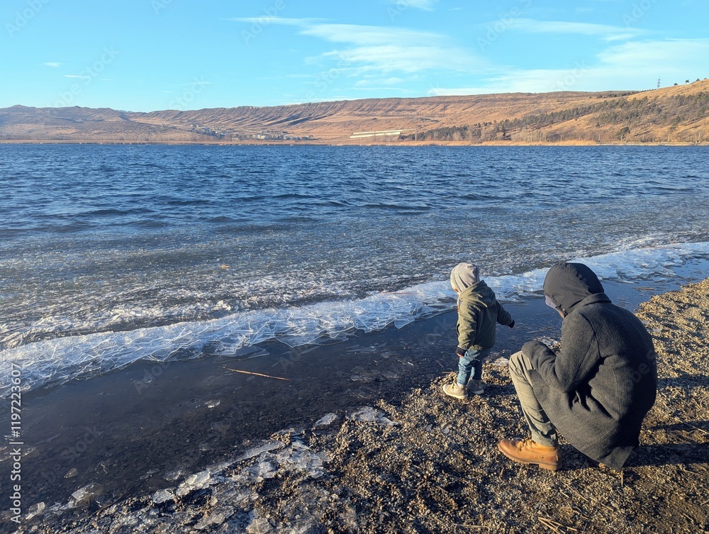 Obraz premium A father and his two-year-old son share a joyful moment playing at the frozen shoreline of a mountain lake on a winter day. Snow surrounds them, creating a serene and picturesque scene of family conne