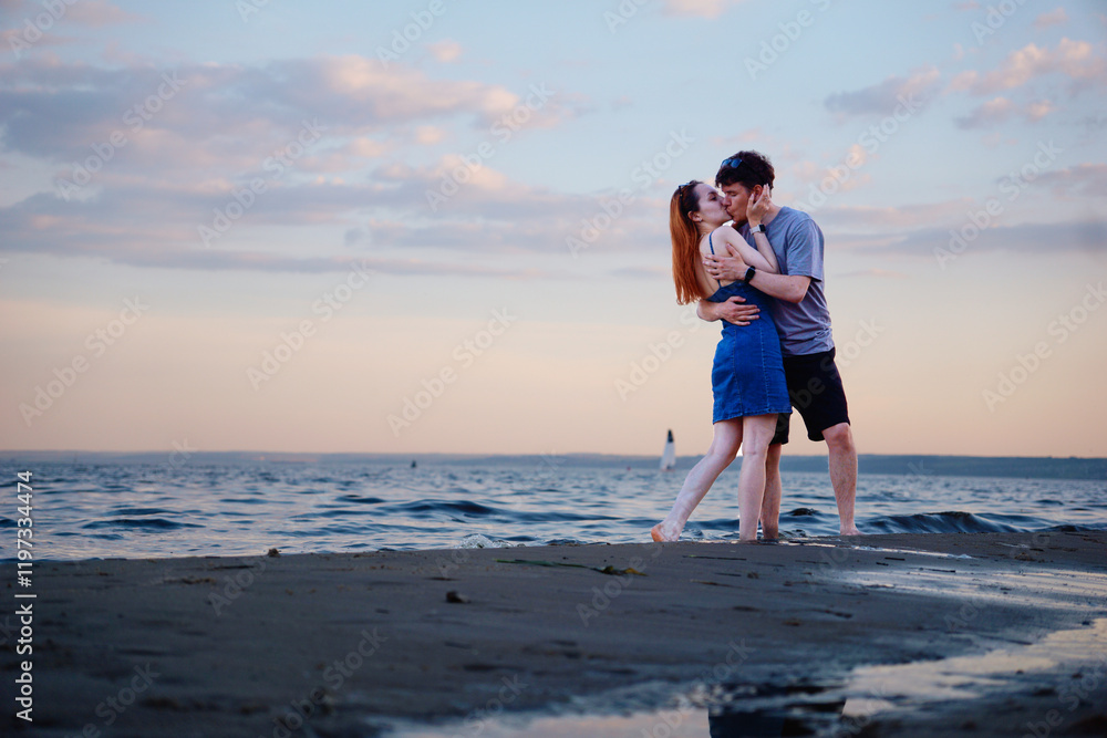 A young couple hugs and kisses on the beach against the backdrop of a beautiful sunset and a passing sailing yacht. Love, date, young man and woman on summer vacation on honeymoon A young couple hugs and kisses on the beach against the backdrop of a beautiful sunset and a passing sailing yacht. Love, date, young man and woman on summer vacation on honeymoon