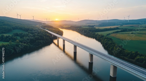 Wallpaper Mural A scenic aerial view of a bridge over a river, surrounded by lush green hills and wind turbines, illuminated by a beautiful sunset. Torontodigital.ca