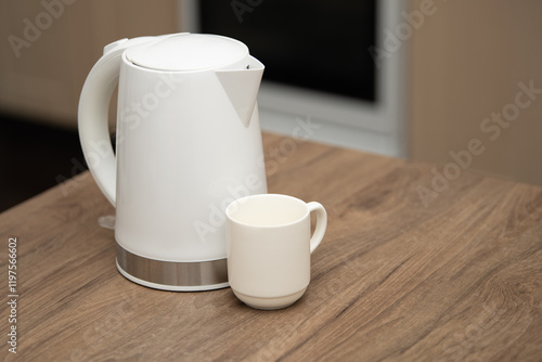 Water is poured from a white electric kettle into a white cup on a wooden table in a cozy kitchen environment