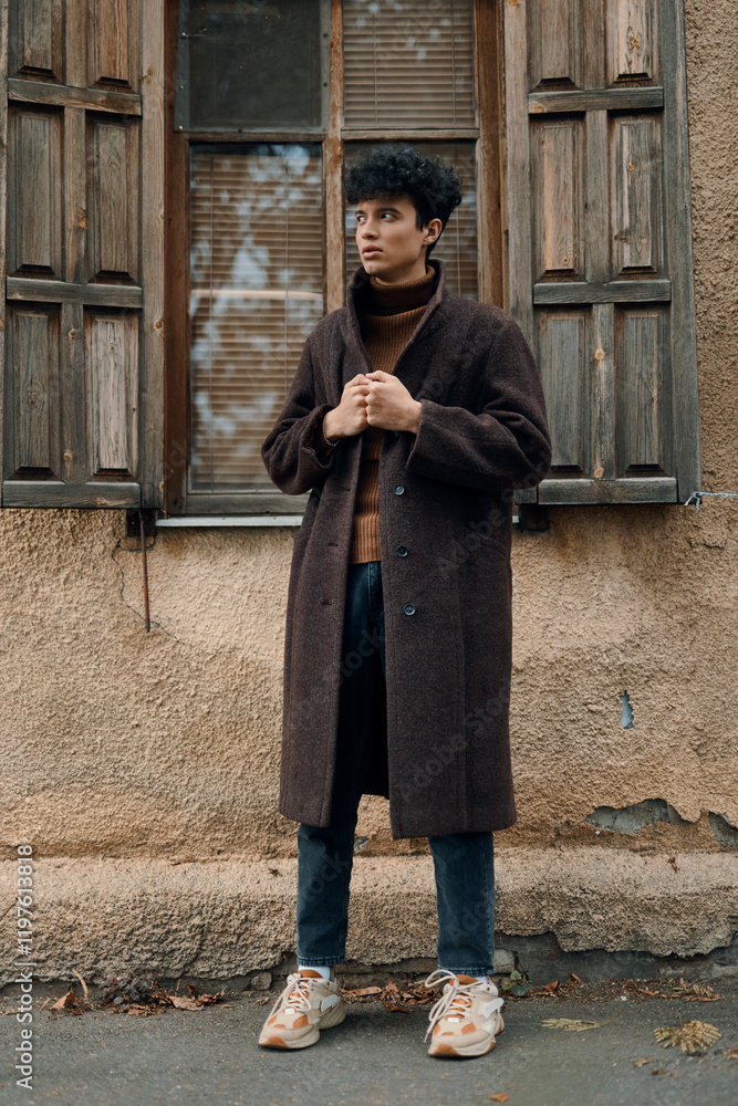 Young man in brown coat and turtleneck sweater standing in front of open window of old building Young man in brown coat and turtleneck sweater standing in front of open window of old building