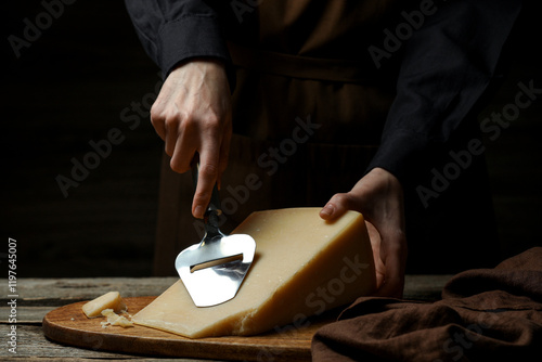 Bilde på lerret Woman cutting delicious cheese with slicer at wooden table against black backgro