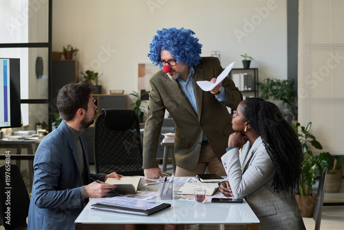 Confident male boss in clown wig and nose looking at one of young subordinates at working meeting in office photo