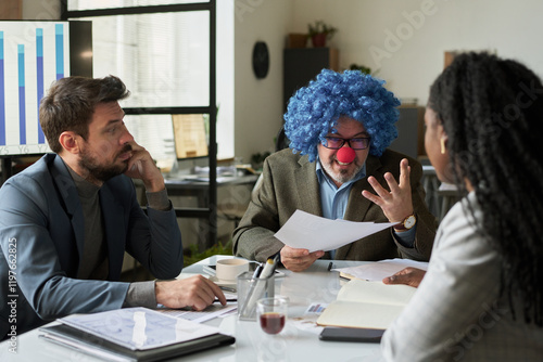 Smiling boss in clown wig looking at one of young subordinates at meeting while explaining points of financial documents photo