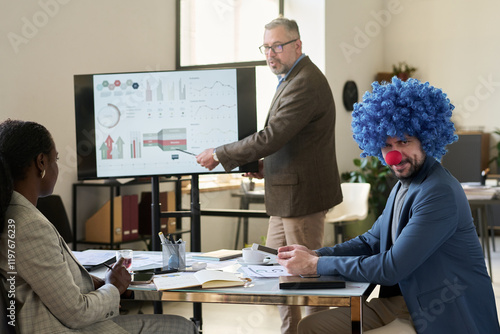 Young smiling businessman in clown wig looking at camera while sitting by workplace against mature boss making presentation photo