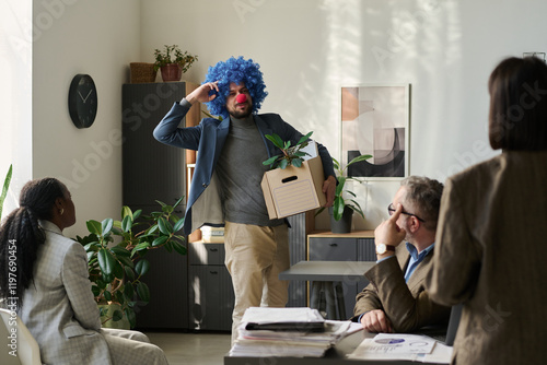 Young male clown employee with box looking at group of colleagues and keeping fingers by head while leaving office photo