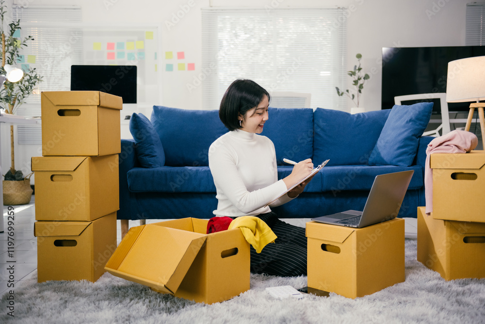 Entrepreneur is smiling while writing on a clipboard and checking inventory on a laptop, surrounded by cardboard boxes full of her products Entrepreneur is smiling while writing on a clipboard and checking inventory on a laptop, surrounded by cardboard boxes full of her products