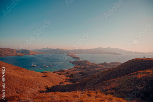 Fototapeta Naklejka Na Ścianę i Meble -  Stunning panoramic view of Labuan Bajo, Indonesia, featuring golden rolling hills, a tranquil blue ocean with anchored sailboats, and a vibrant sunset glow. 
