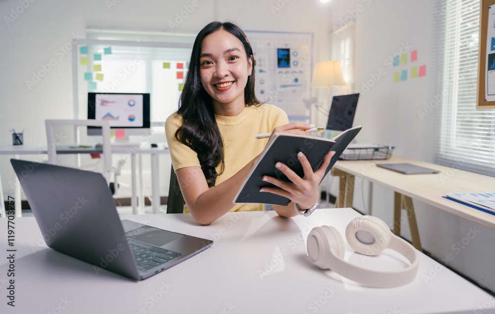 Young asian businesswoman happily takes notes at her modern office desk, surrounded by technology and natural light Young asian businesswoman happily takes notes at her modern office desk, surrounded by technology and natural light