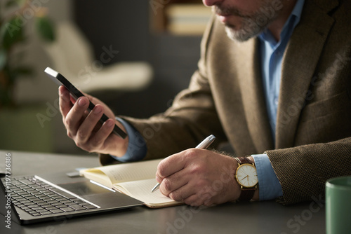 Hands of mature male boss in formalwear sitting by workplace, texting in mobile phone and making notes in notebook photo