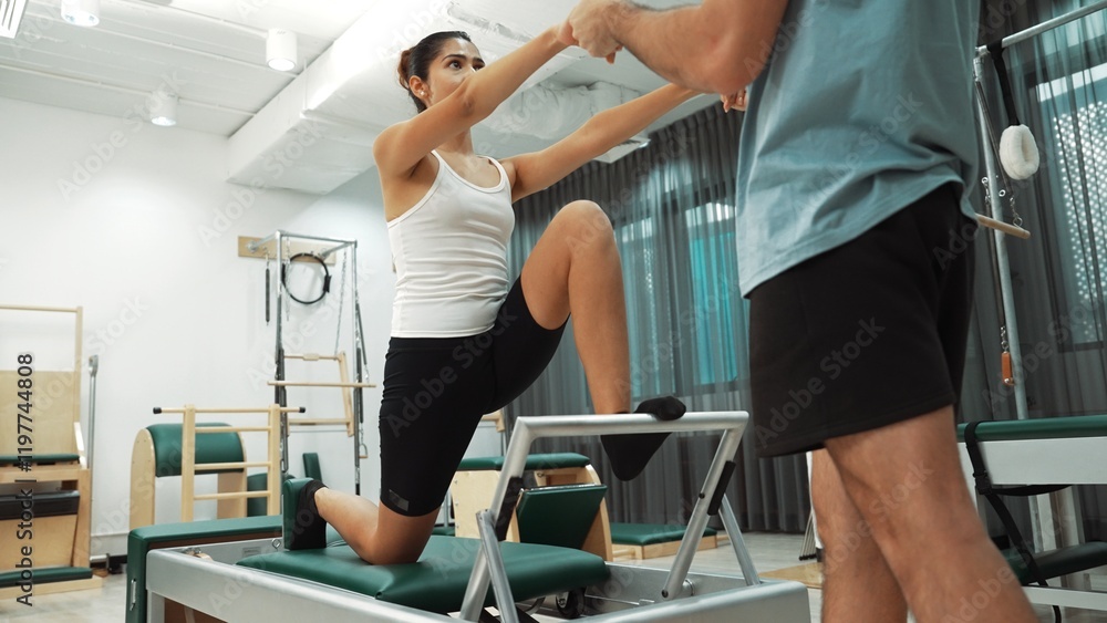 Woman receiving assistance in split stretch exercise during pilates class on trapeze table for flexibility and core training. Student doing split stretch on reformer for improving balance. Habituate. Woman receiving assistance in split stretch exercise during pilates class on trapeze table for flexibility and core training. Student doing split stretch on reformer for improving balance. Habituate.