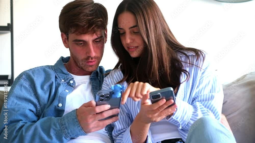 Two people in their early 30s, casually dressed, sit close to each other in a home setting, scrolling through social media content on their phones, particularly watching videos. Two people in their early 30s, casually dressed, sit close to each other in a home setting, scrolling through social media content on their phones, particularly watching videos.