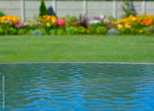 Obraz na płótnie niebo odbite w szklanym blacie stołu na tle ogrodu z trawnikiem i kwiatami, sky reflected in the glass table against the background of garden with flowers, blue sky reflected in glass table, iluzja w salonie