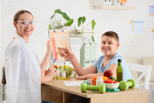 Overweight boy and nutritionist with meal planner at table in clinic. Diet co...