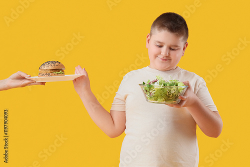 Overweight boy with vegetable salad rejecting burger on yellow background. Di...