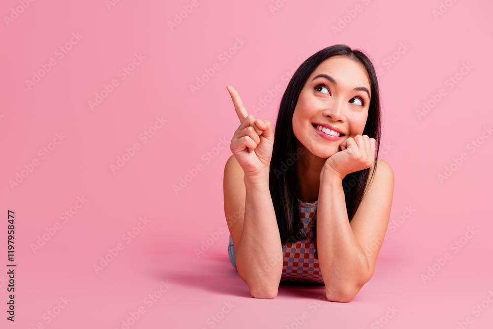Joyful young woman pointing upward with a smile on a pink background, showcasing her charm and style Joyful young woman pointing upward with a smile on a pink background, showcasing her charm and style