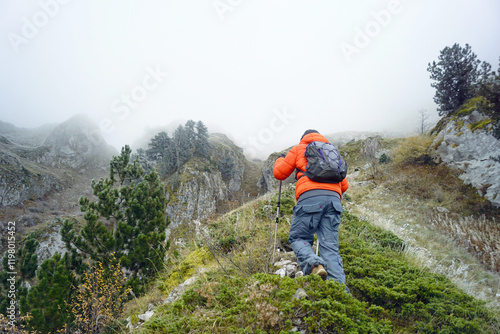 Prokletije National Park trails: A tourist during a challenging route in the Montenegrin mountains. Photographed from behind, a man climbs up a steep slope using hiking poles on a cold, foggy day photo