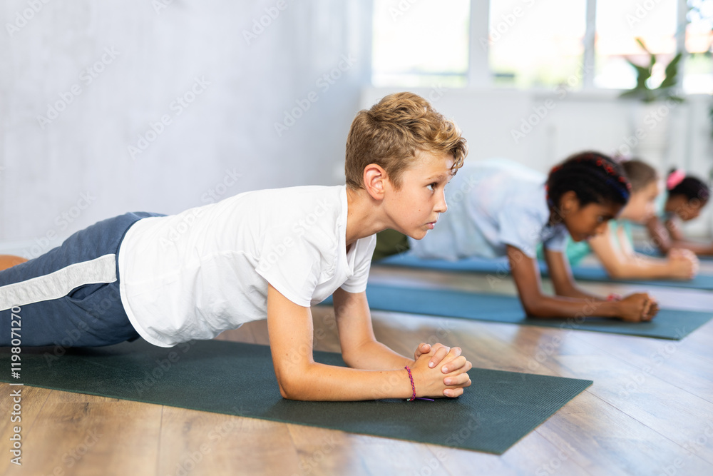 In fitness studio with group of friends, boy children perform plank pose. Male teen participates in yoga practice. Concept of active lifestyle of youthful generation In fitness studio with group of friends, boy children perform plank pose. Male teen participates in yoga practice. Concept of active lifestyle of youthful generation