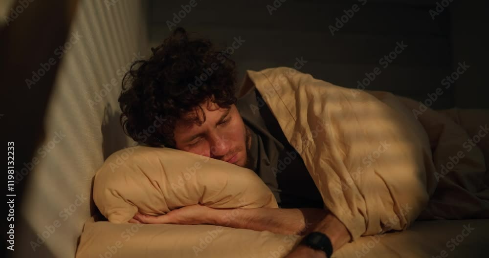 A sleepy man with curly hair wakes up in the morning of his bed and looks at the phone screen at the alarm clock in the morning A sleepy man with curly hair wakes up in the morning of his bed and looks at the phone screen at the alarm clock in the morning