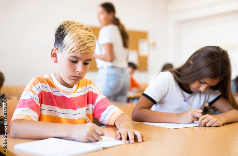 Portrait of concentrated tween boy studying in classroom, listening to schoolteacher and writing in notebook Portrait of concentrated tween boy studying in classroom, listening to schoolteacher and writing in notebook