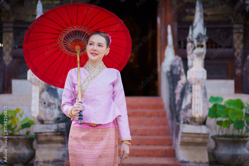 Fototapeta premium Beautiful woman portrait in fashion Lanna style in traditional Thai dress, wearing an elegant pink dress and holding a red umbrella, in Wat Ton Kwen Temple in Chiang Mai, Thailand.