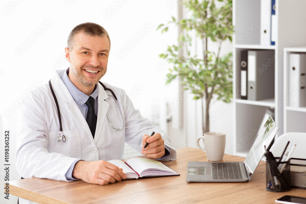 Fototapeta premium Professional Help Concept. Portrait of happy handsome male doctor in stethoscope and white unifrom sitting at table with laptop at his office, posing, looking at camera and writing in his notebook
