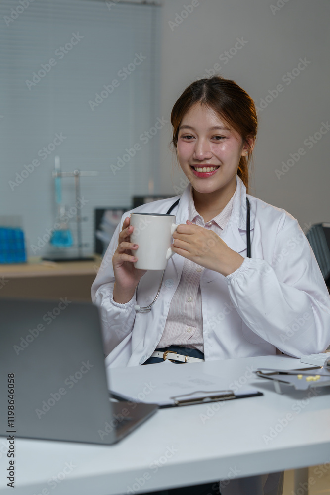 Young female asian doctor wearing lab coat and stethoscope, drinking from a white mug while working on a laptop, attending an online telehealth consultation in her bright office Young female asian doctor wearing lab coat and stethoscope, drinking from a white mug while working on a laptop, attending an online telehealth consultation in her bright office